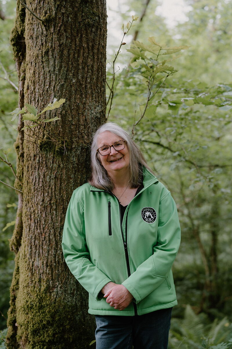 A woman with grey hair and glasses smiles beside a large tree trunk in woodland, wearing a green jacket with a round embroidered badge.