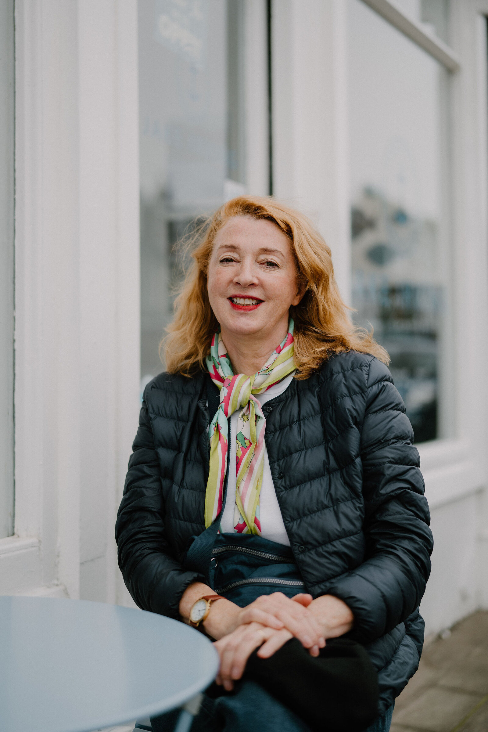 A woman in a black puffer jacket and floral scarf smiles at an outdoor café table, a shopfront window behind her.