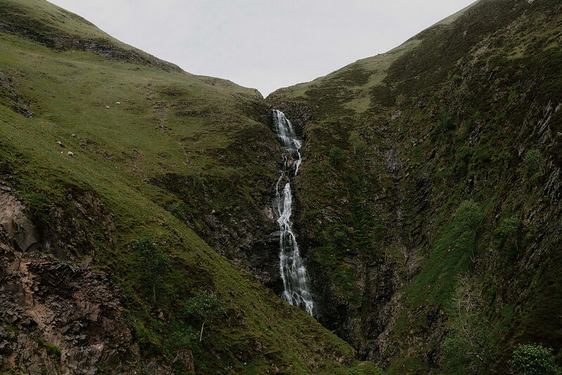 A waterfall drops through a narrow green gully between two grassy hills, the white water catching the light.