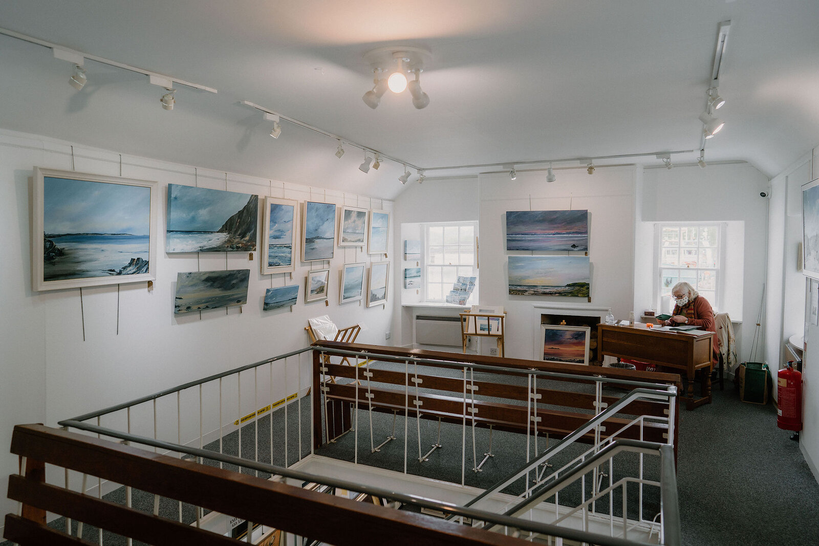 Framed coastal landscape paintings line the walls of an upper gallery, a metal railing overlooking a person working at a desk below.