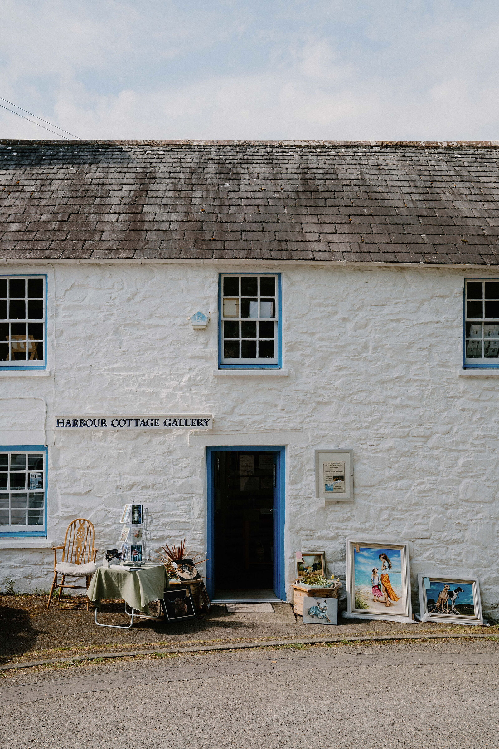 A sign reading "HARBOUR COTTAGE GALLERY" is set into a whitewashed building's blue-trimmed wall, framed paintings and a folding table displayed outside the open door.
