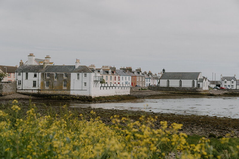 Whitewashed and pastel-coloured houses line a harbour shore, yellow wildflowers filling the grassy foreground.
