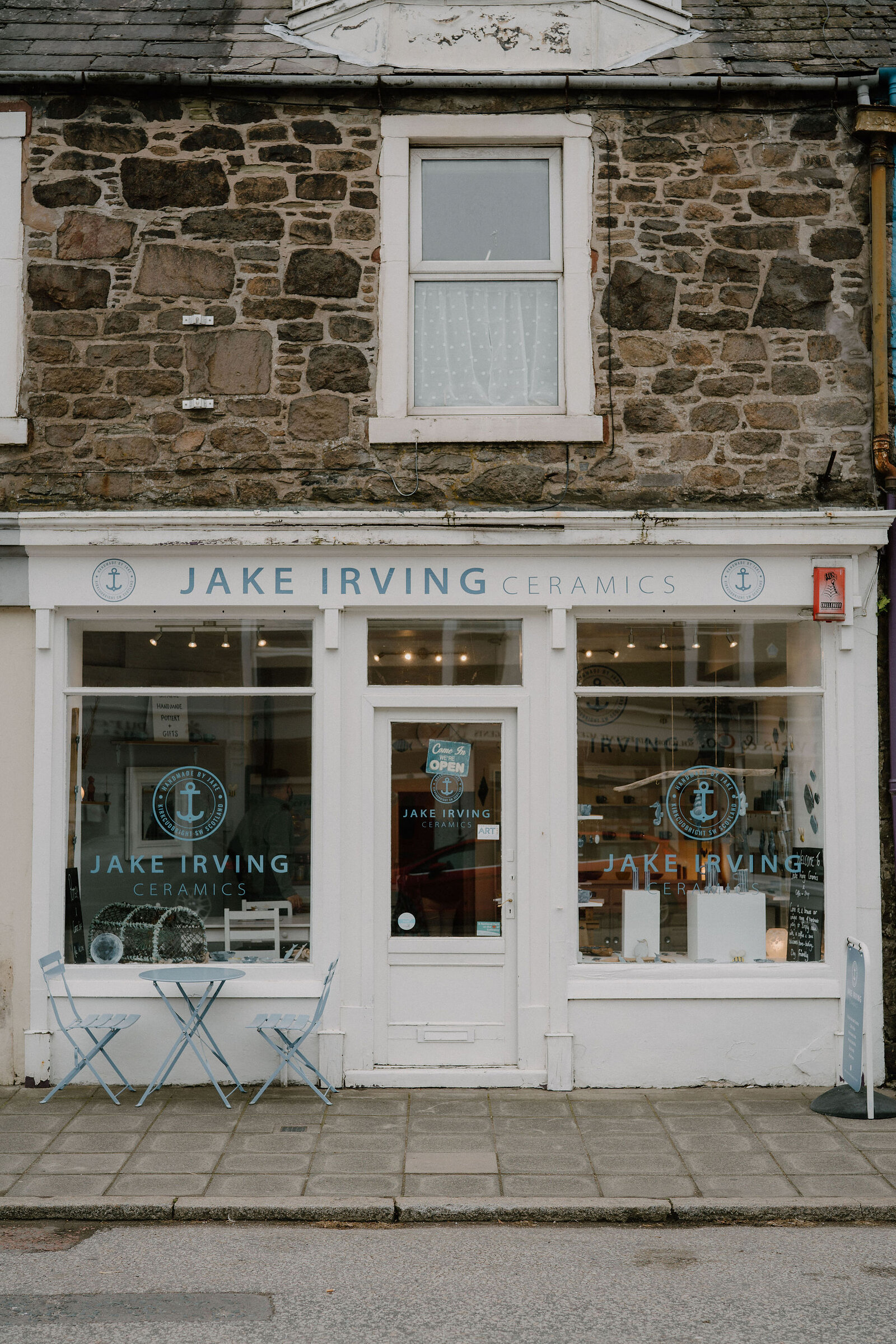 A white shopfront reads "JAKE IRVING CERAMICS" beside an anchor logo, a blue bistro table and chairs set outside beneath the window.