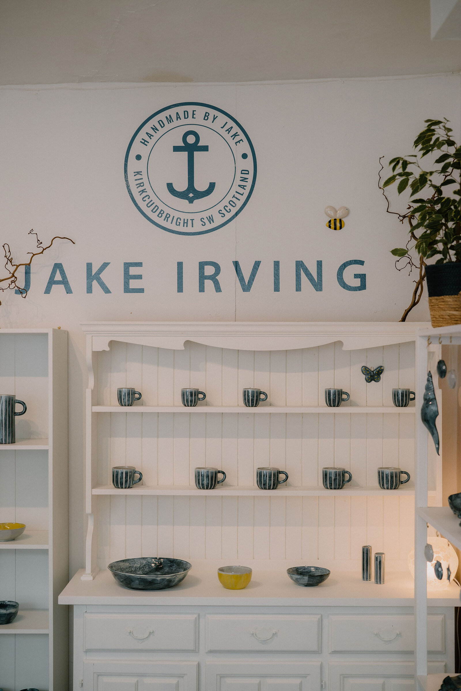 A logo reading "JAKE IRVING — HANDMADE BY JAKE, KIRKCUDBRIGHT SW SCOTLAND" with an anchor motif is painted above white shelving stocked with striped mugs and bowls.