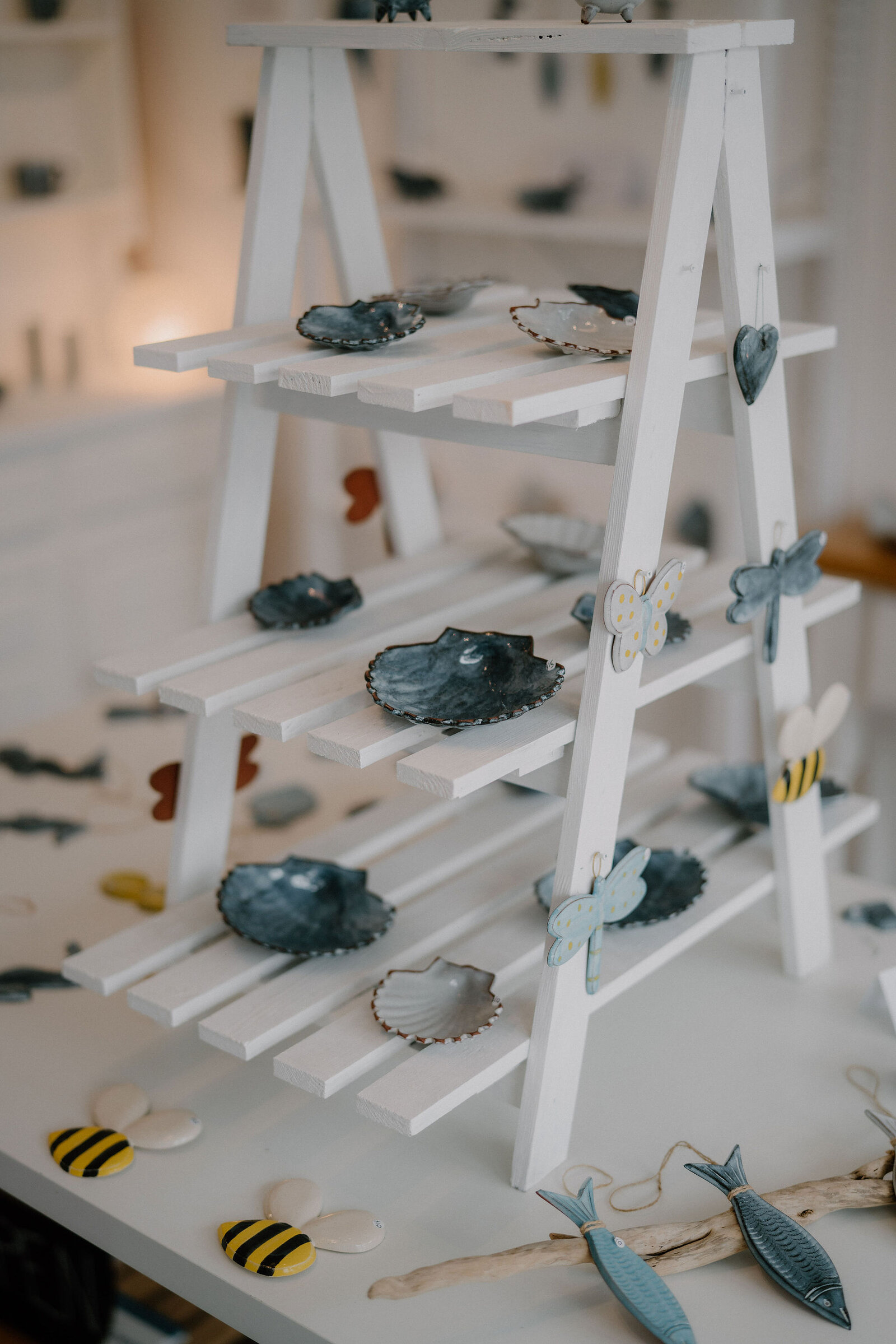 A white wooden display ladder holds ceramic shell-shaped dishes, butterfly and bee ornaments arranged among small driftwood fish figures.