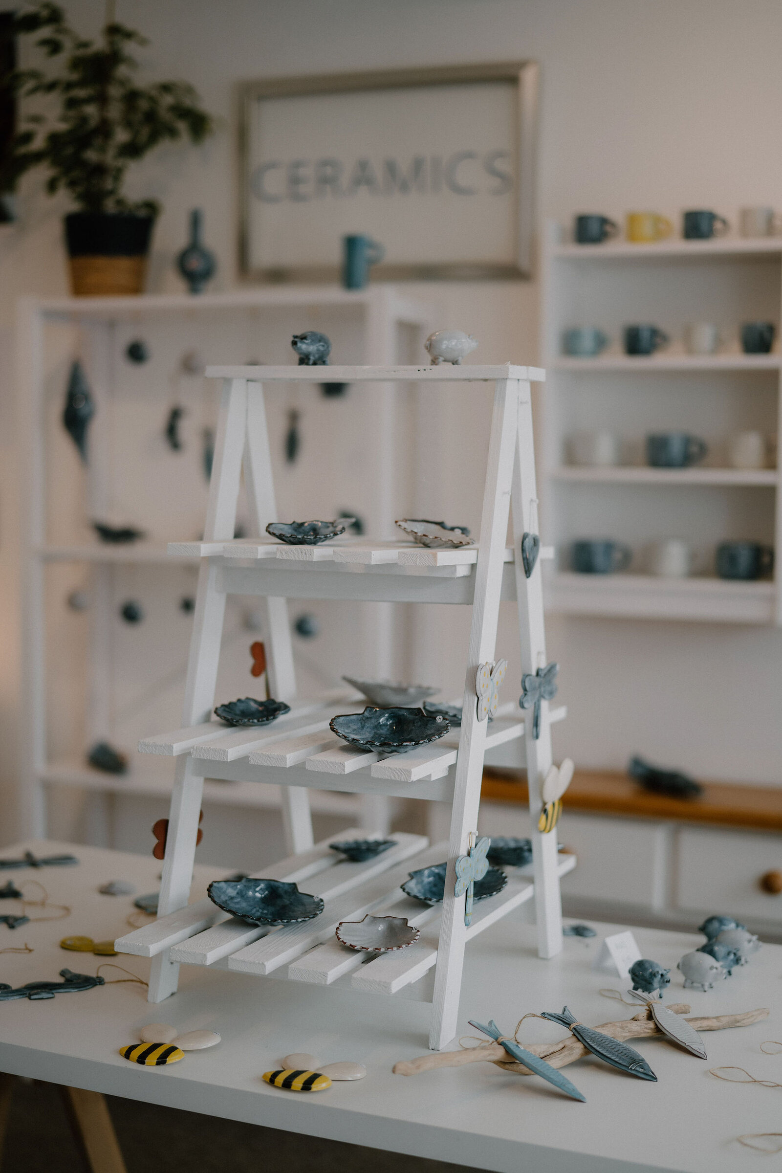 A white display ladder holds ceramic dishes and small ornaments beneath a sign reading "CERAMICS", shelves of blue and white mugs standing behind.