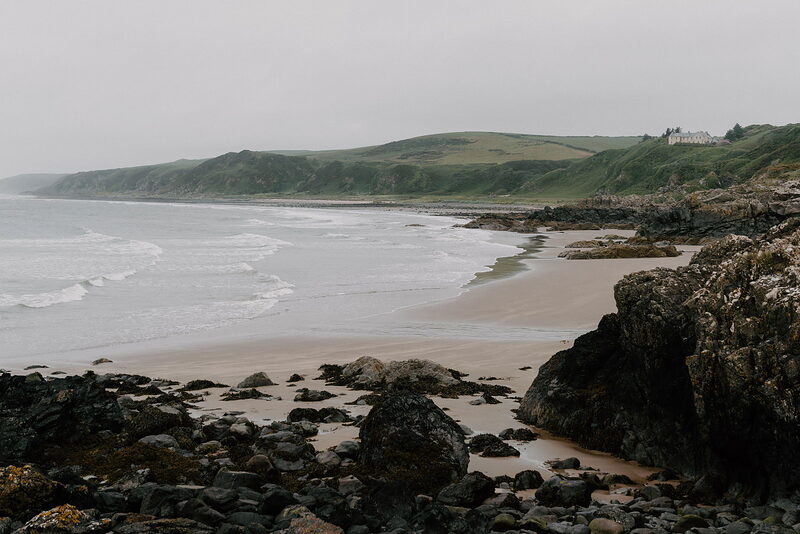 A sandy beach studded with dark rocks curves beneath green hills, waves breaking gently along the shore.