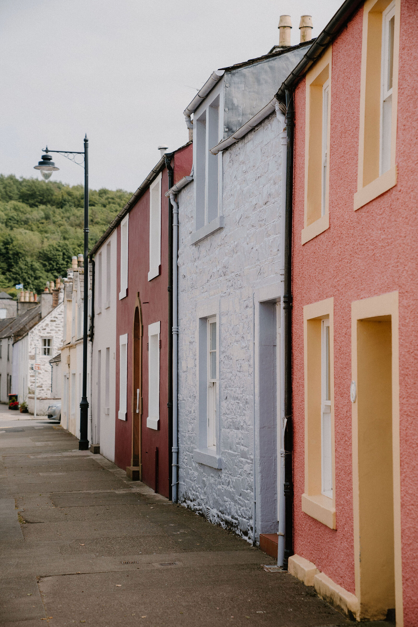 A street of terraced houses painted in maroon, white and pink runs downhill past a wooded slope, a lamppost standing at the corner.
