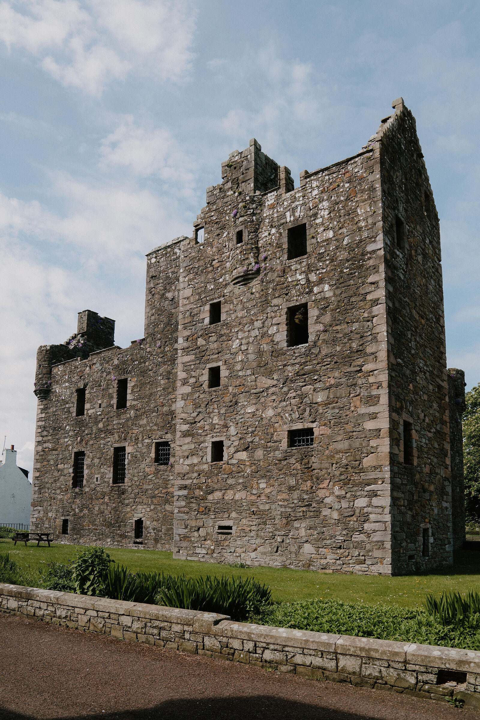 A tall ruined stone mansion with rows of empty window openings and a stepped gable stands behind a low wall, a bench set on the lawn beside it.