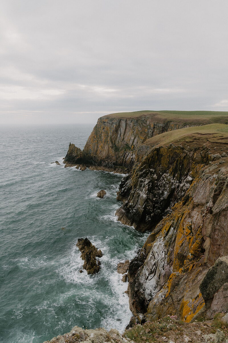 Layered rock cliffs streaked with orange lichen drop to a churning sea, a jagged sea stack rising near the shore.