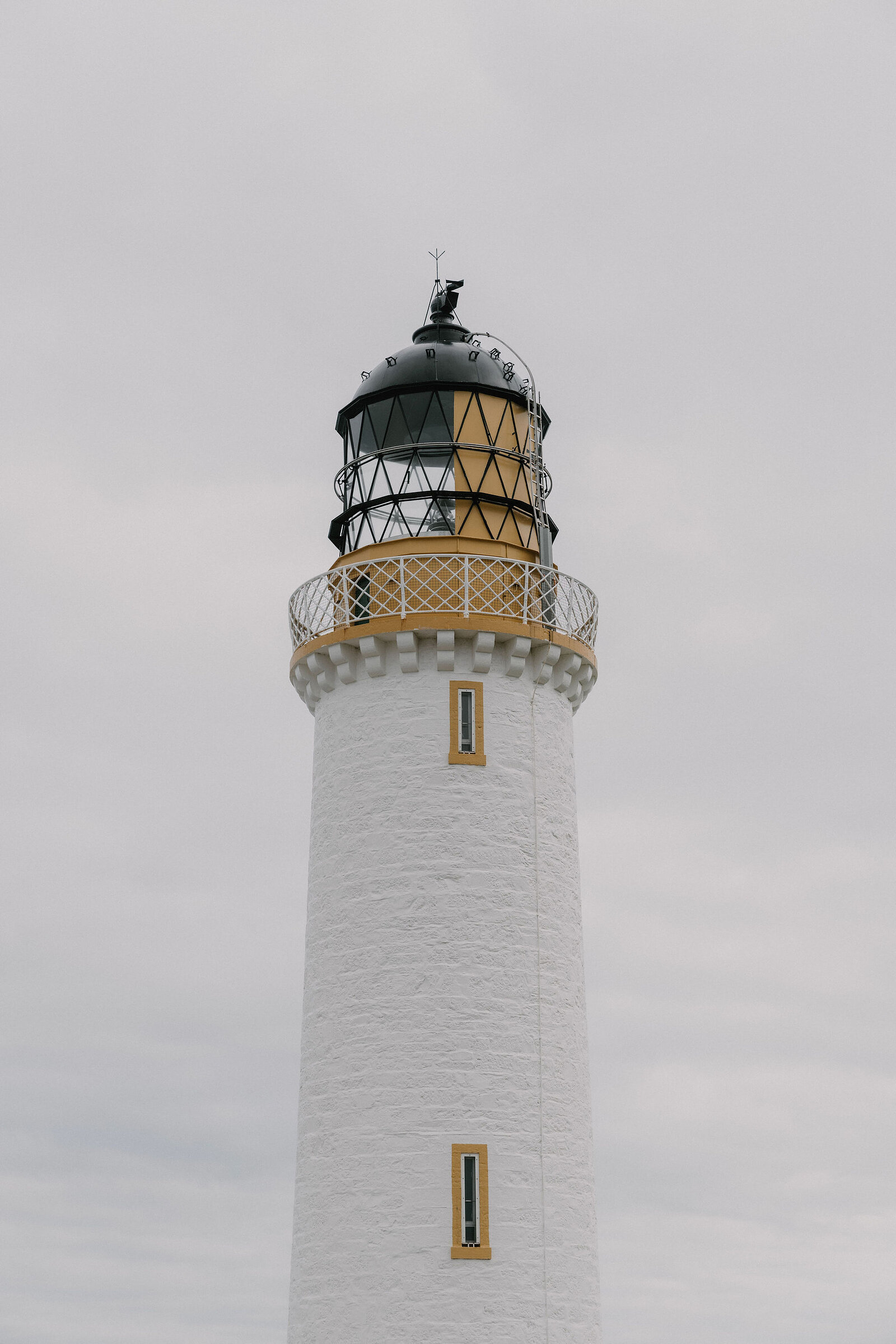 The upper section of a white lighthouse shows its glazed lantern room and gallery railing against an overcast sky.