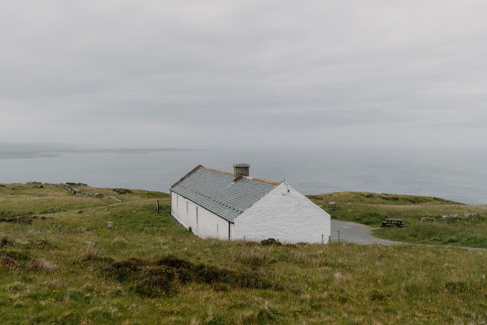 A long white farm building with a slate roof stands on a grassy clifftop, the sea stretching hazily into the distance behind.