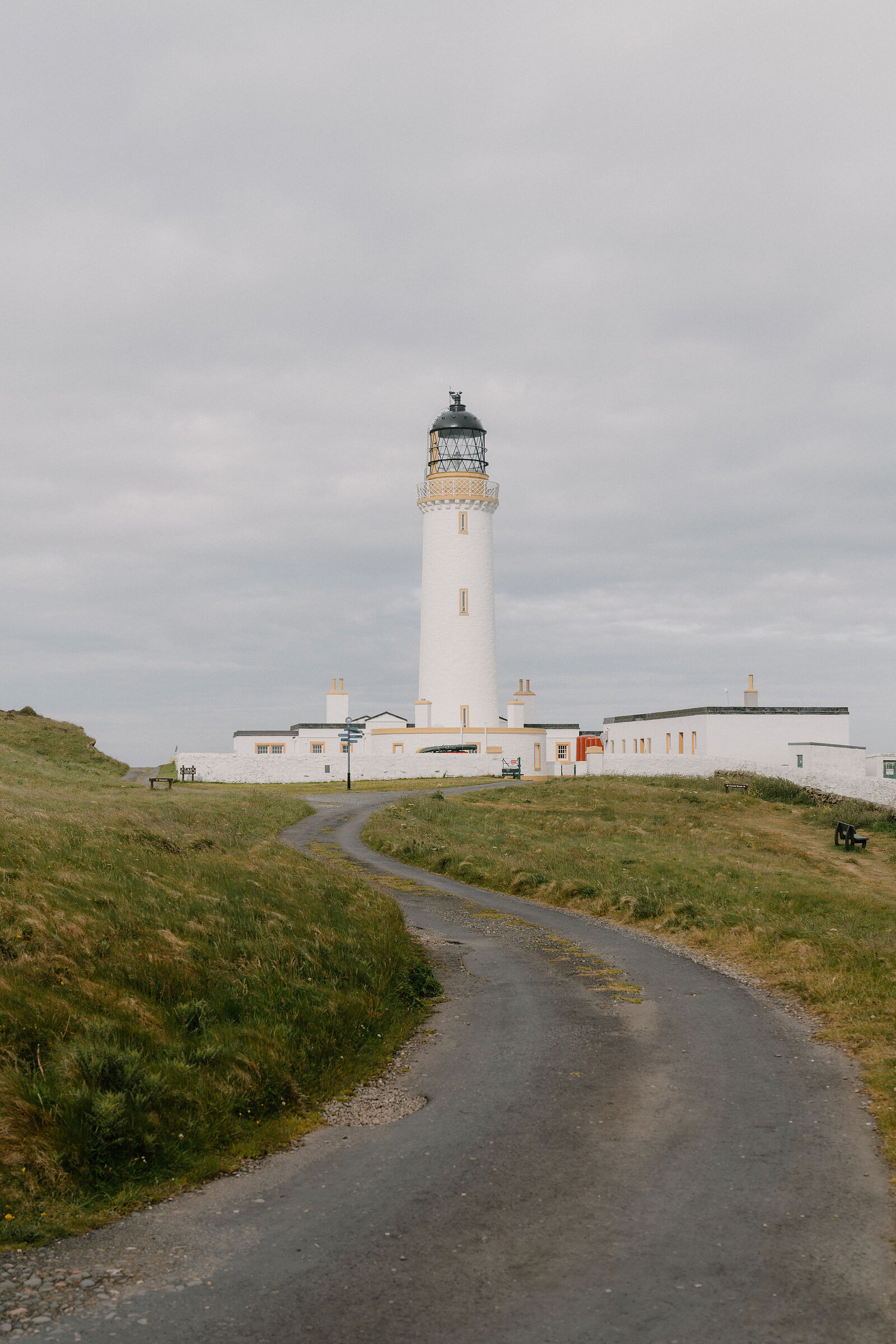 A tarmac path curves up a grassy headland towards a tall white lighthouse and its outbuildings.