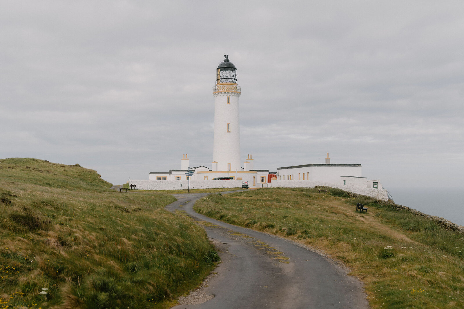 A tarmac path winds up a grassy headland towards a tall white lighthouse and its outbuildings, the sea visible beyond.