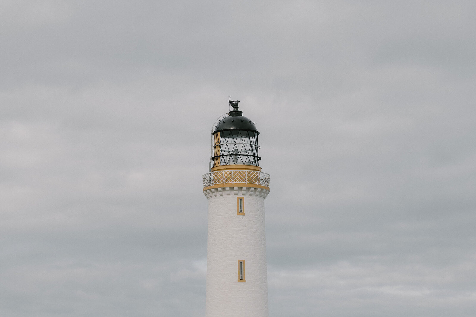 The black lantern room and gallery of a white lighthouse stand out against an overcast sky.