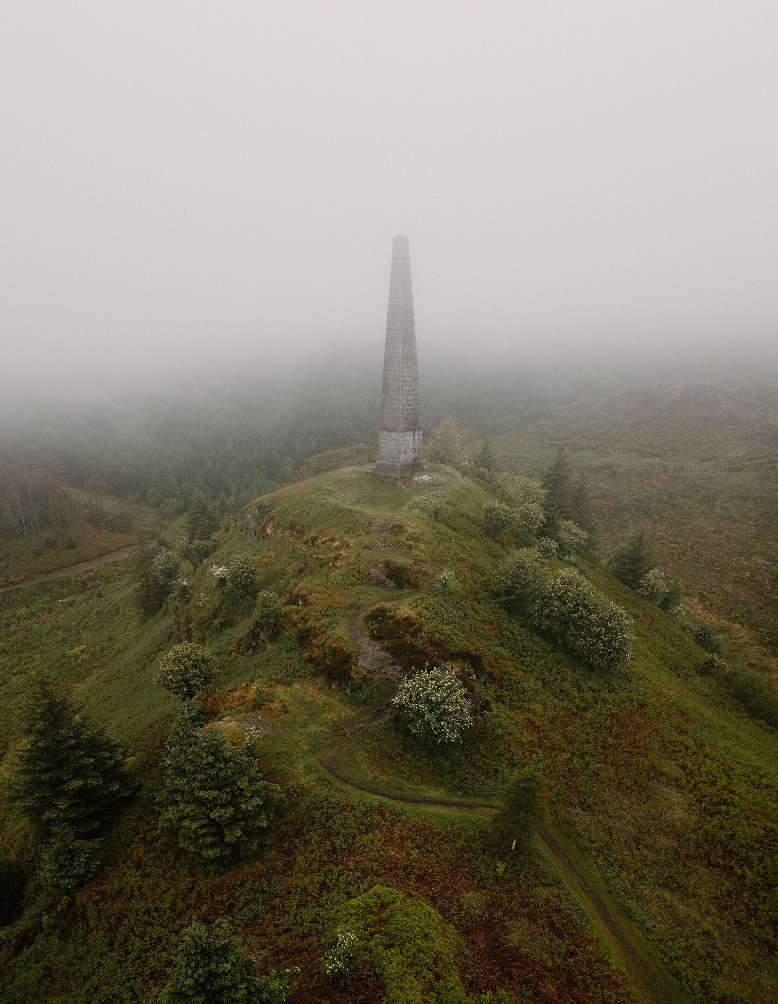 An aerial view shows a stone obelisk standing on a hilltop wrapped in mist, hawthorn bushes and a winding path covering the slopes below.