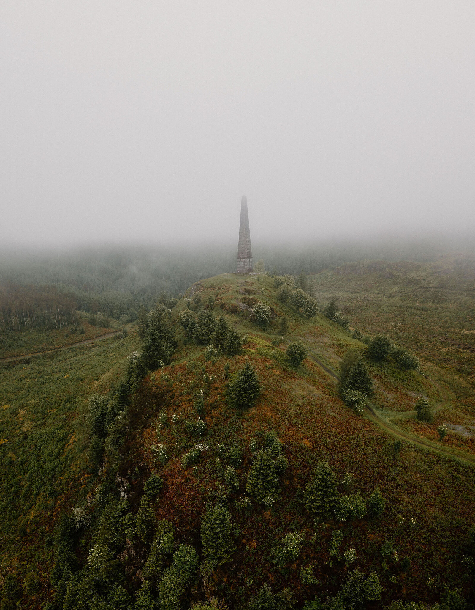 An aerial view shows a stone obelisk on a misty hilltop surrounded by autumn-coloured moorland and scattered pine trees.