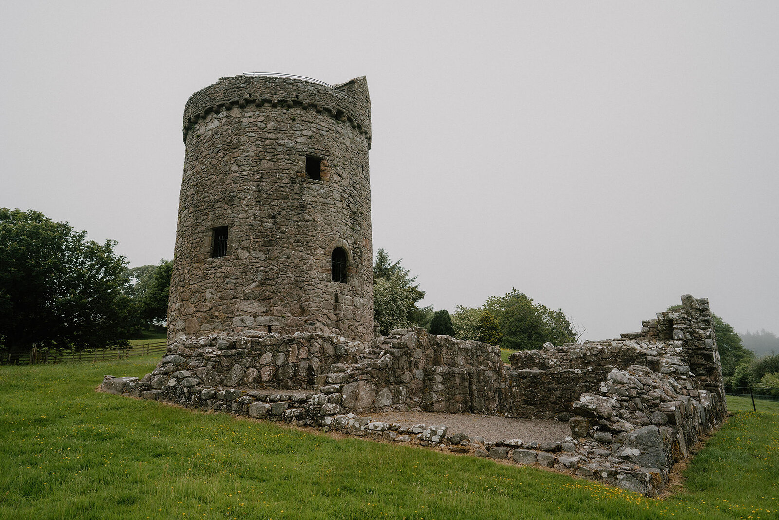 A round stone tower with a corbelled top stands within the low ruined walls of a courtyard, trees lining the field behind.