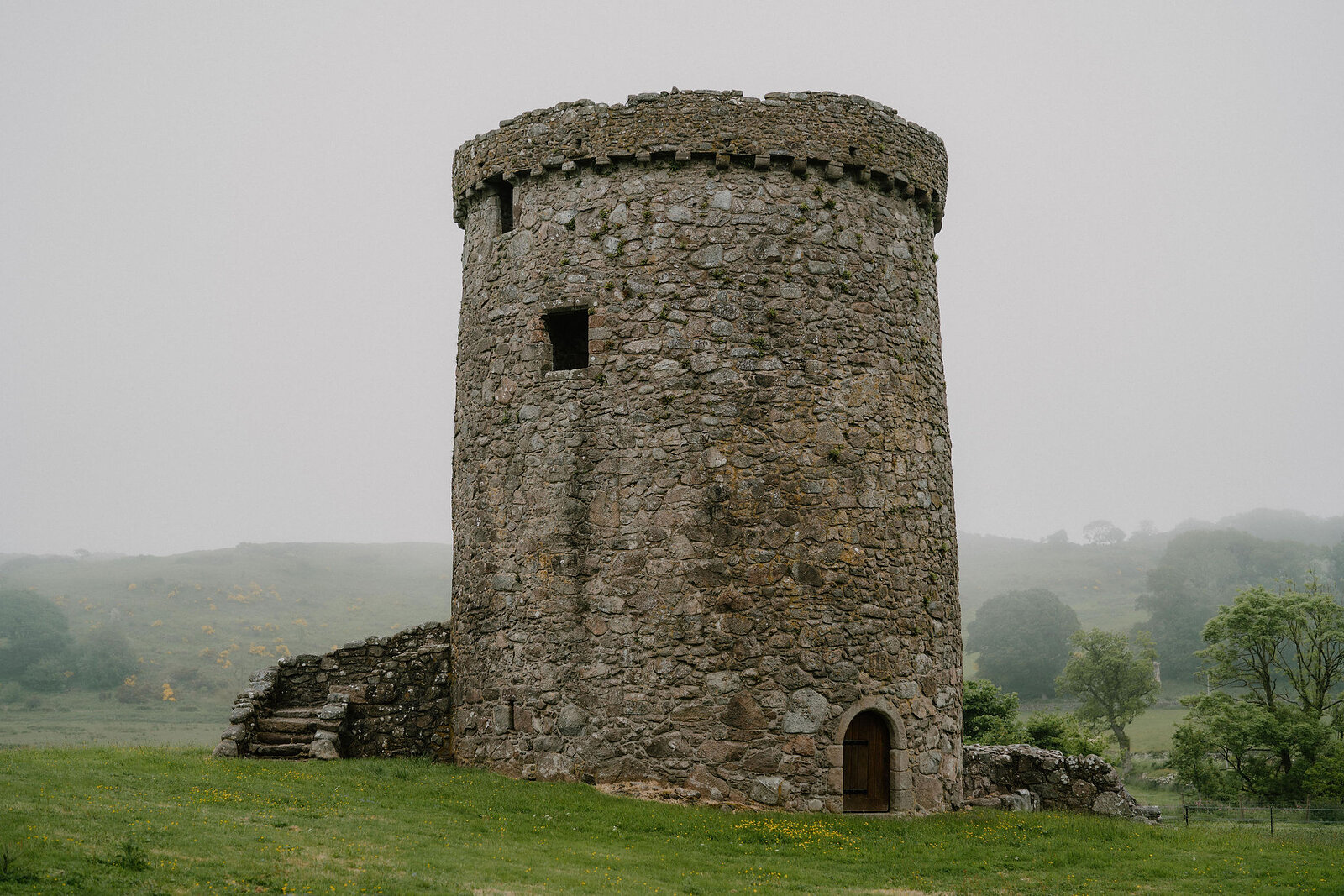 A round stone tower with a corbelled top and small square opening stands in a field, low ruined walls extending from its base.