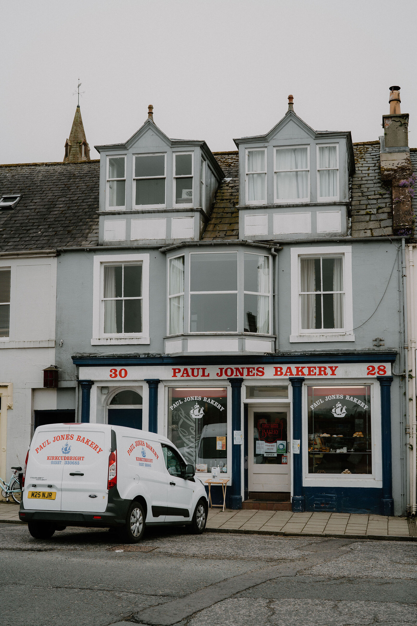 A tall stone building with bay-windowed dormers is signed "Paul Jones Bakery" in red lettering, a branded van parked outside its blue-trimmed shopfront.