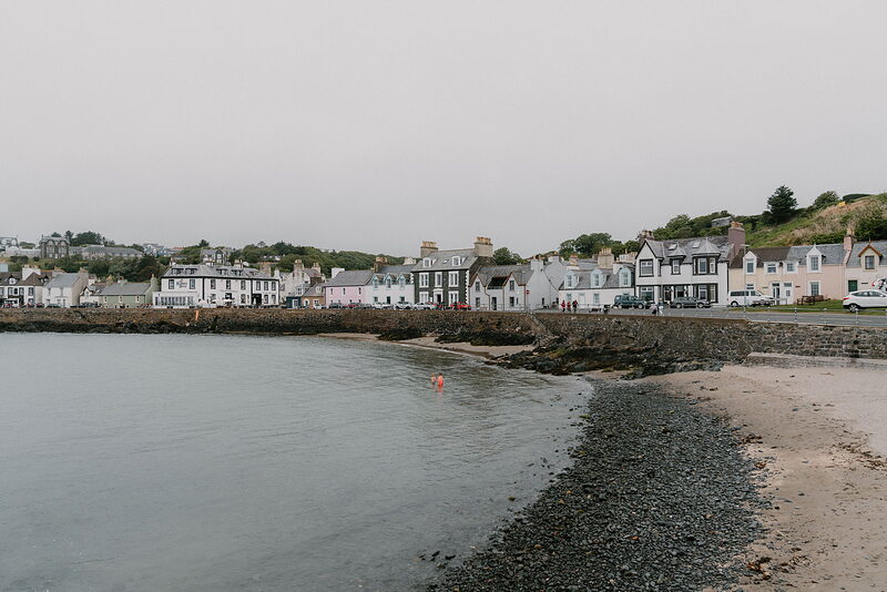 A row of colourful houses lines a curving bay, a pebble beach sloping down to calm water.
