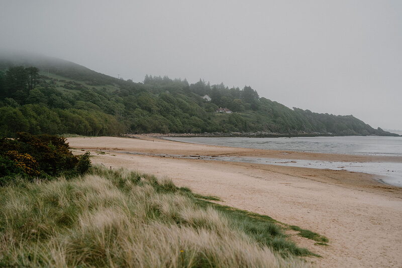 A wide sandy beach curves beneath a wooded, mist-covered hillside, gorse bushes growing along the dune edge.