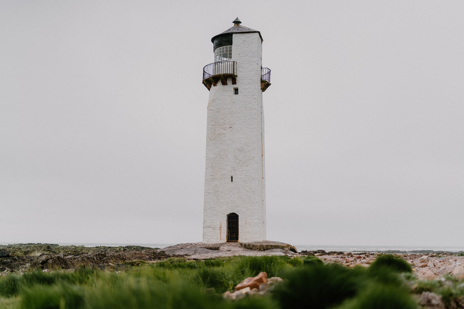 A white lighthouse with a black lantern room stands on a rocky shore, seaweed-covered rocks and grass filling the foreground.