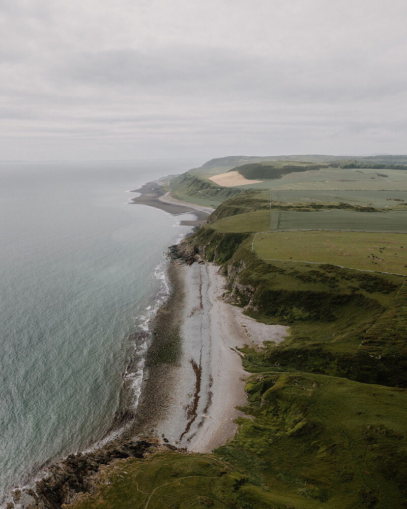An aerial view shows layered coastal cliffs curving along the sea, a sandy beach and green farmland stretching inland.