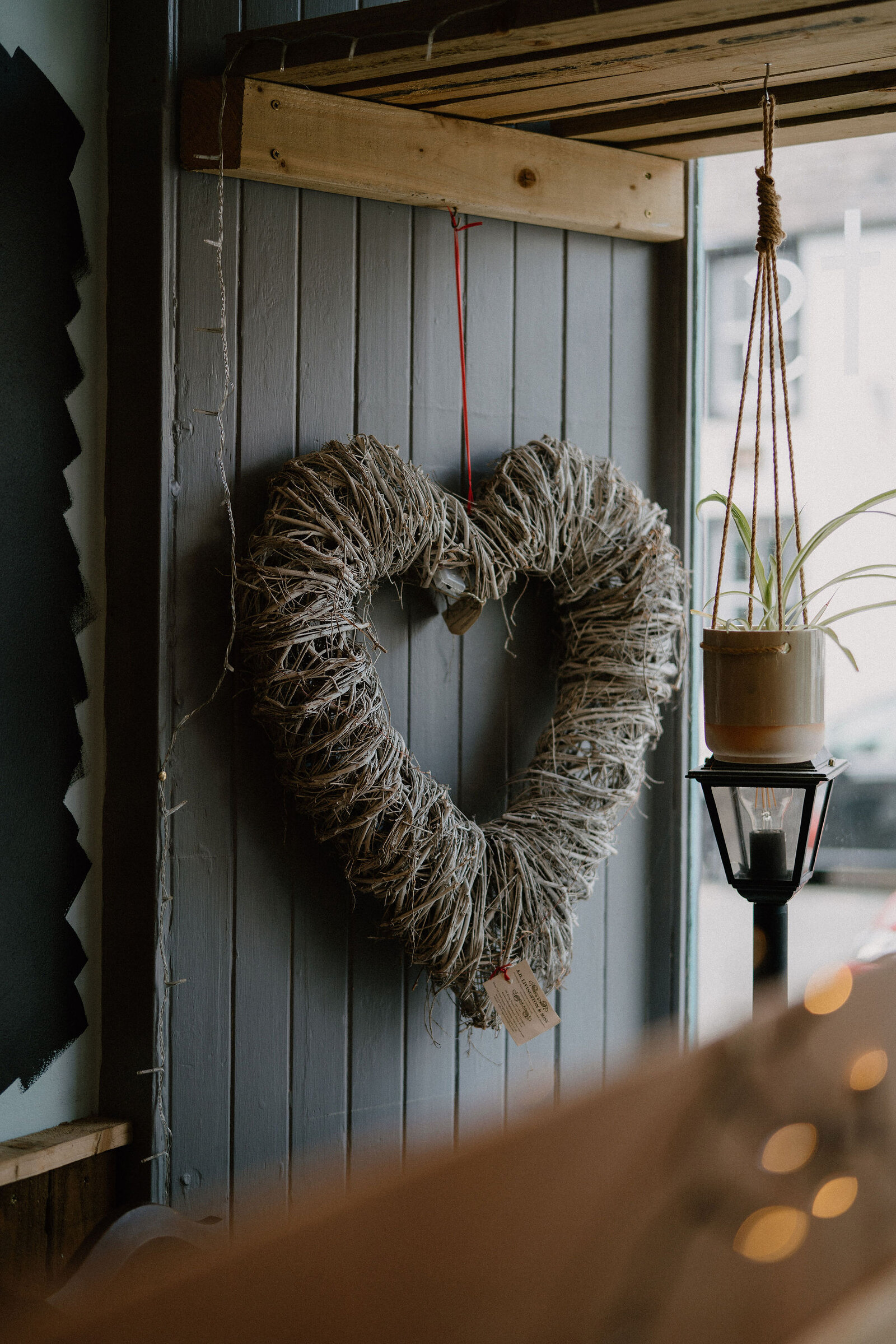 A wreath of twisted twine shaped like a heart hangs on a panelled wall beside a hanging basket plant and a lit lantern.