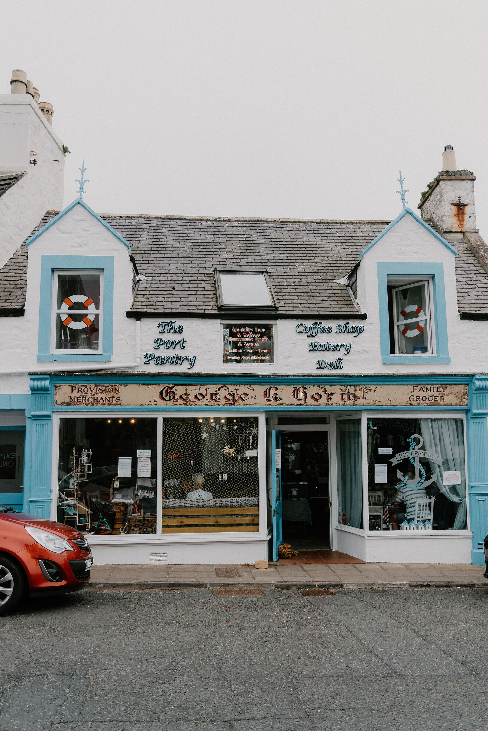 A blue and white harbourside building carries signs reading 'The Port Pantry, Coffee Shop, Bakery, Deli' and 'George & Gow, Provision Merchants, Family Grocer'.