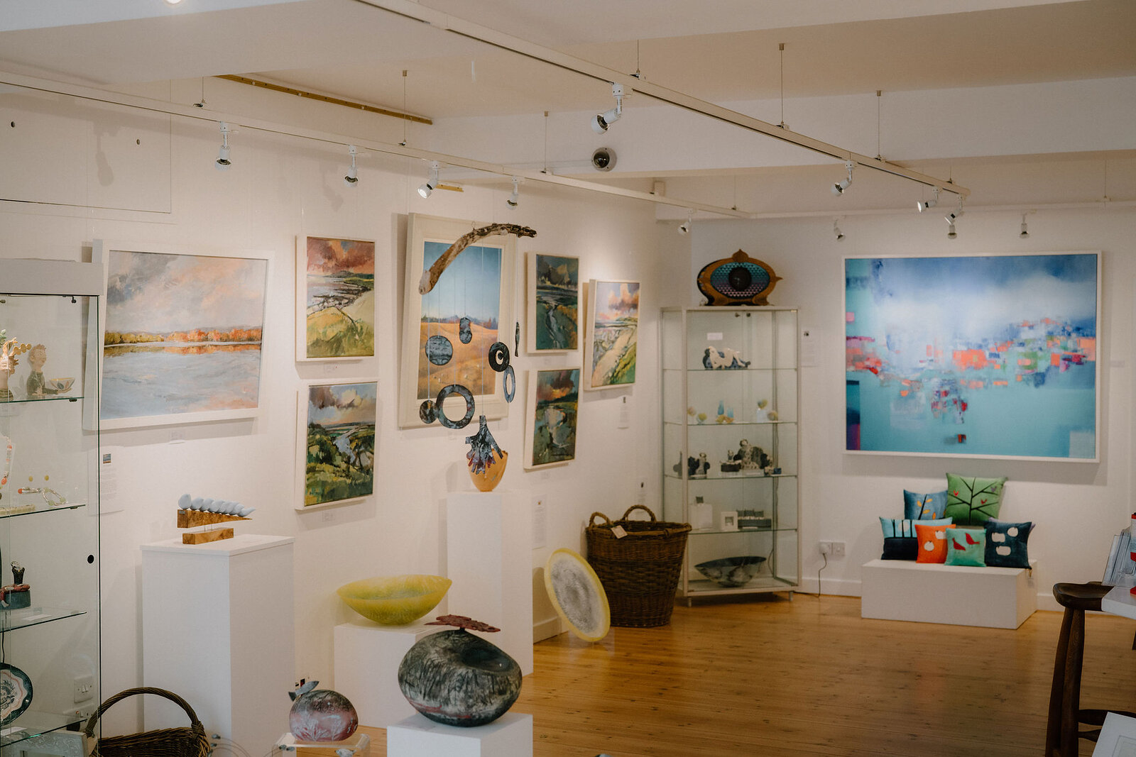 Paintings, ceramics and a woven basket are displayed in a gallery room, glass cabinets of small ornaments standing beside framed coastal scenes.