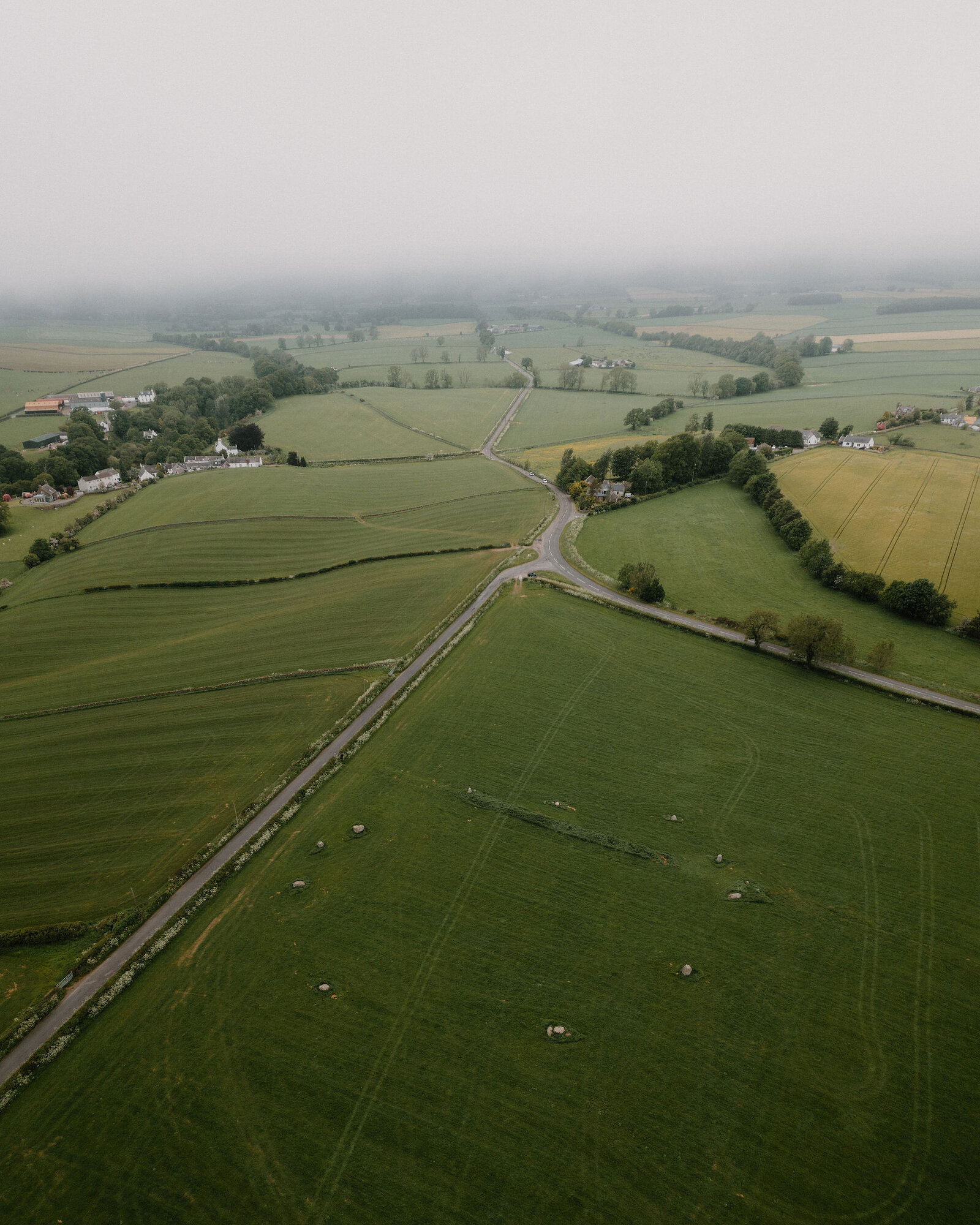 An aerial view over misty farmland shows a crossroads, dry stone walls dividing green and golden fields, and a scattered farmhouse.
