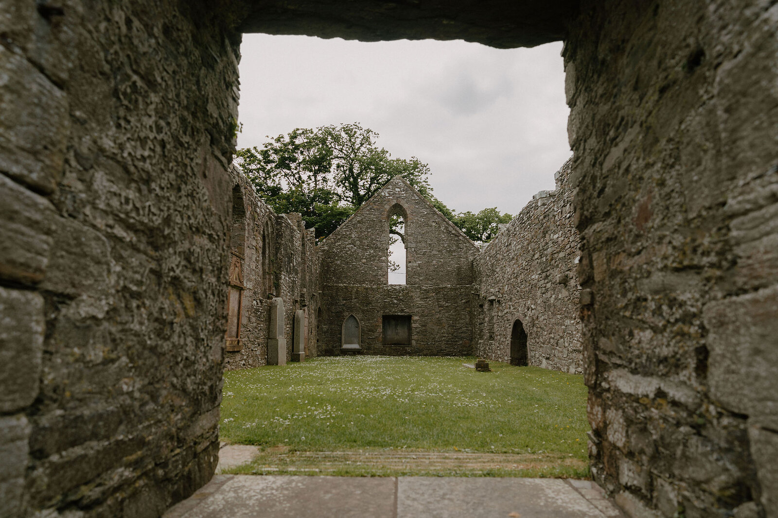 A stone archway frames the roofless nave of a ruined chapel, a tree and lawn visible through the traceried east window.