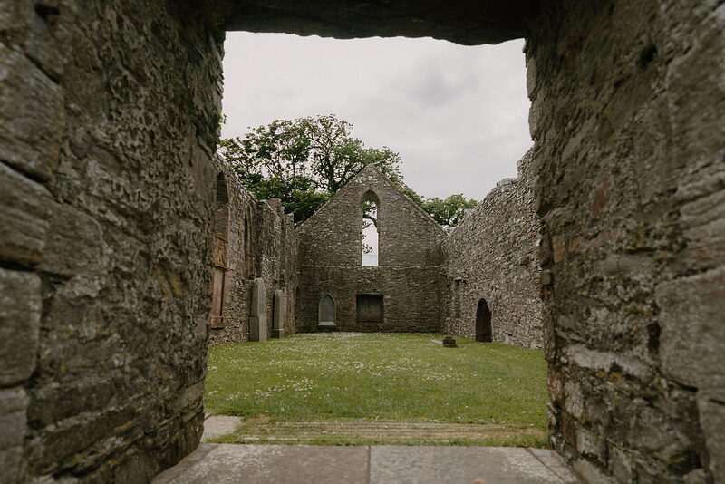 A stone archway frames the roofless nave of a ruined chapel, a tree and lawn visible through the traceried east window.