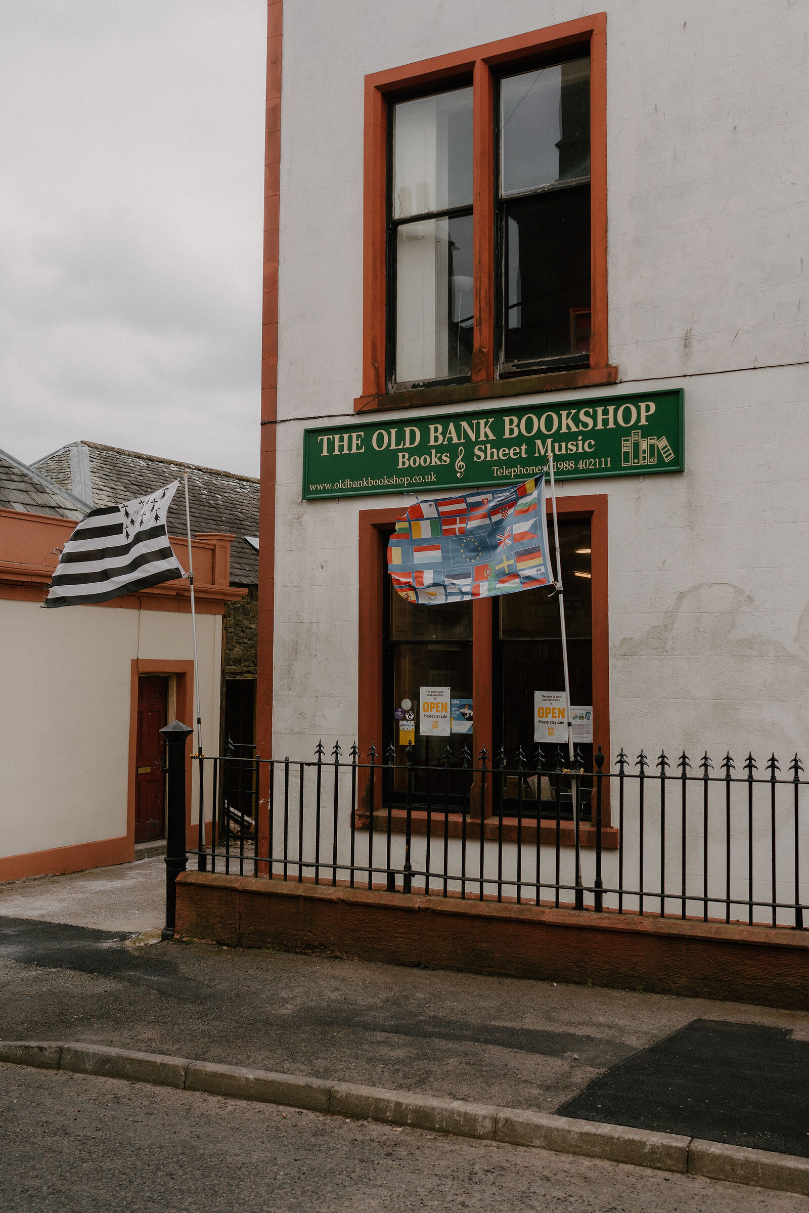 A white building carries a sign reading 'The Old Bank Bookshop, Books & Sheet Music', national flags strung along a railing outside.