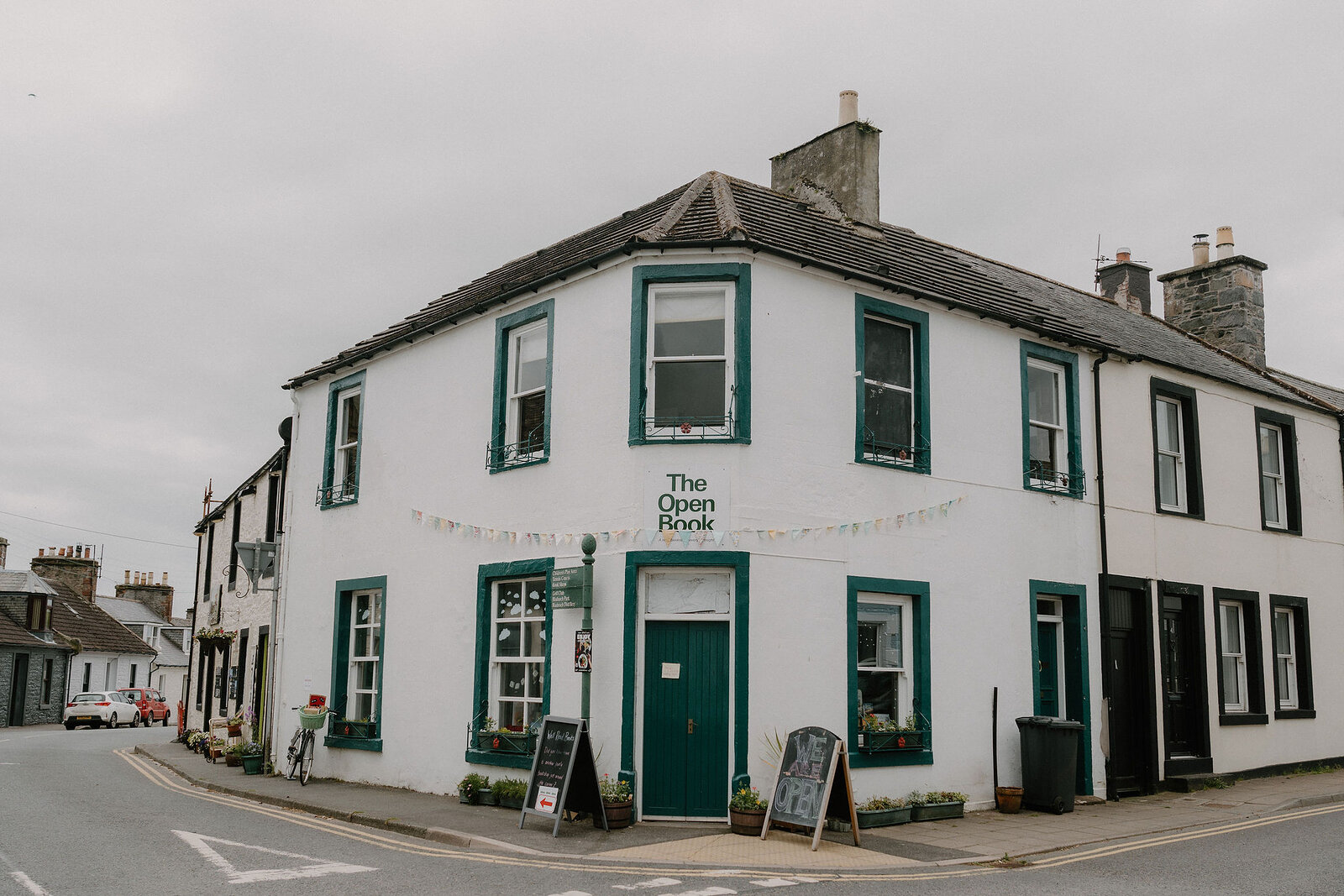 A white corner building with green-painted window frames carries a sign reading 'The Open Book', a chalkboard and bunting outside.