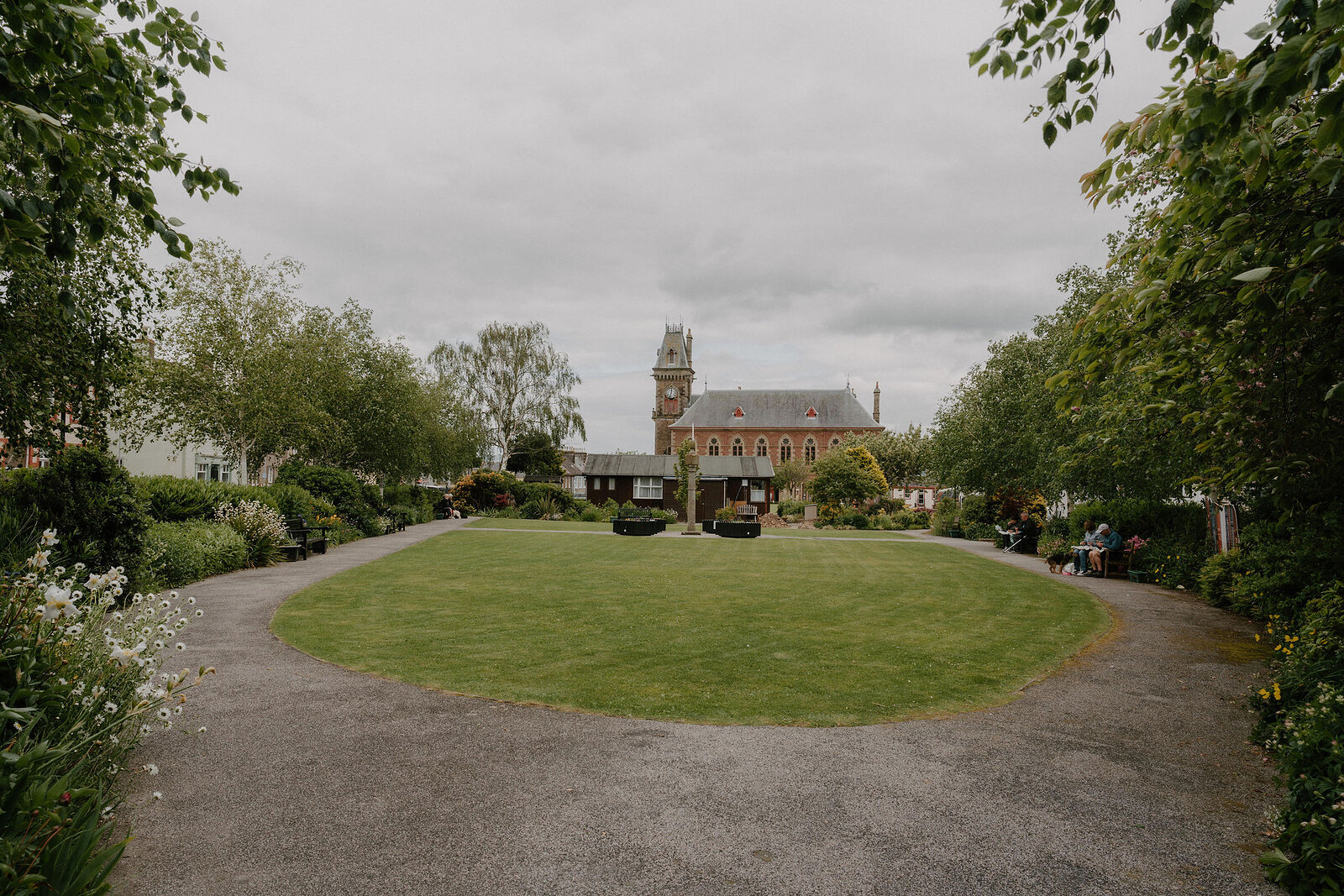 A circular lawn with a war memorial sits in a public garden bordered by hedges, a red-brick clock tower building visible beyond the trees.
