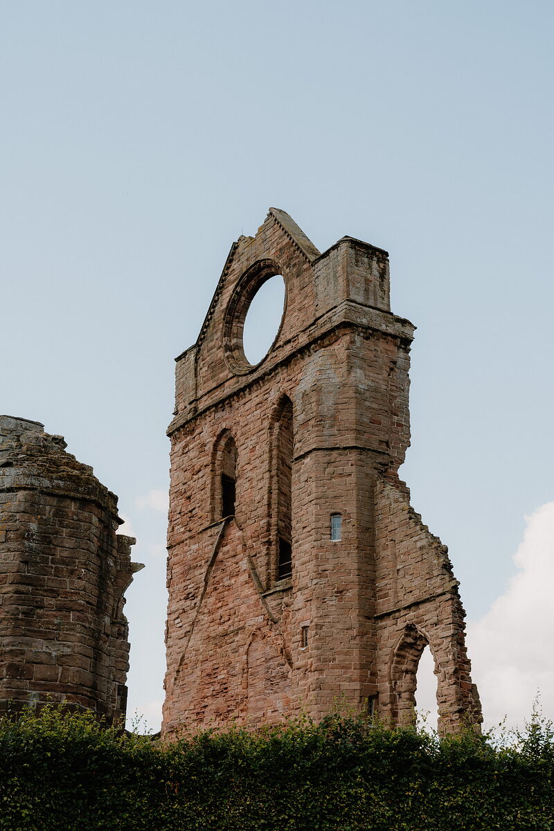 A large circular window pierces the red sandstone gable of a ruined abbey, a smaller tower rising beside it against a clear sky.