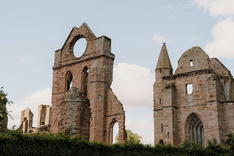 A ruined abbey's round window and a slender pointed tower rise together against a pale sky, more broken walls visible beyond.