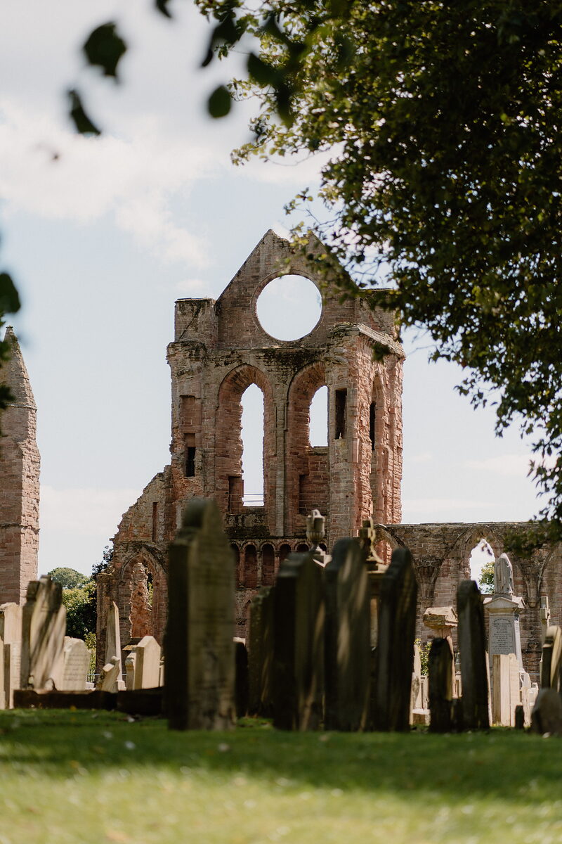 A ruined abbey's round window rises above a churchyard of worn headstones, trees framing the view on both sides.