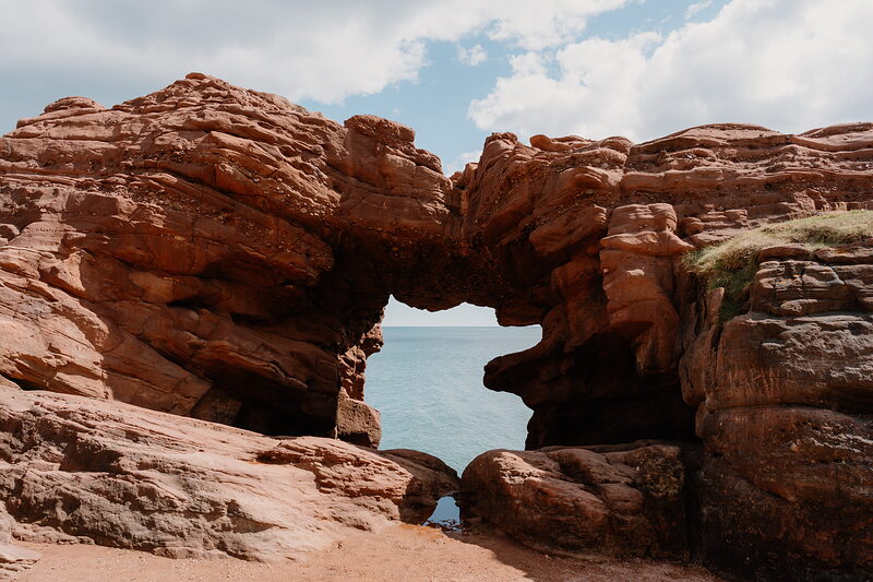 A red sandstone sea arch frames a gap of blue water, a small second opening breaking through the rock below and a sandy beach at its foot.
