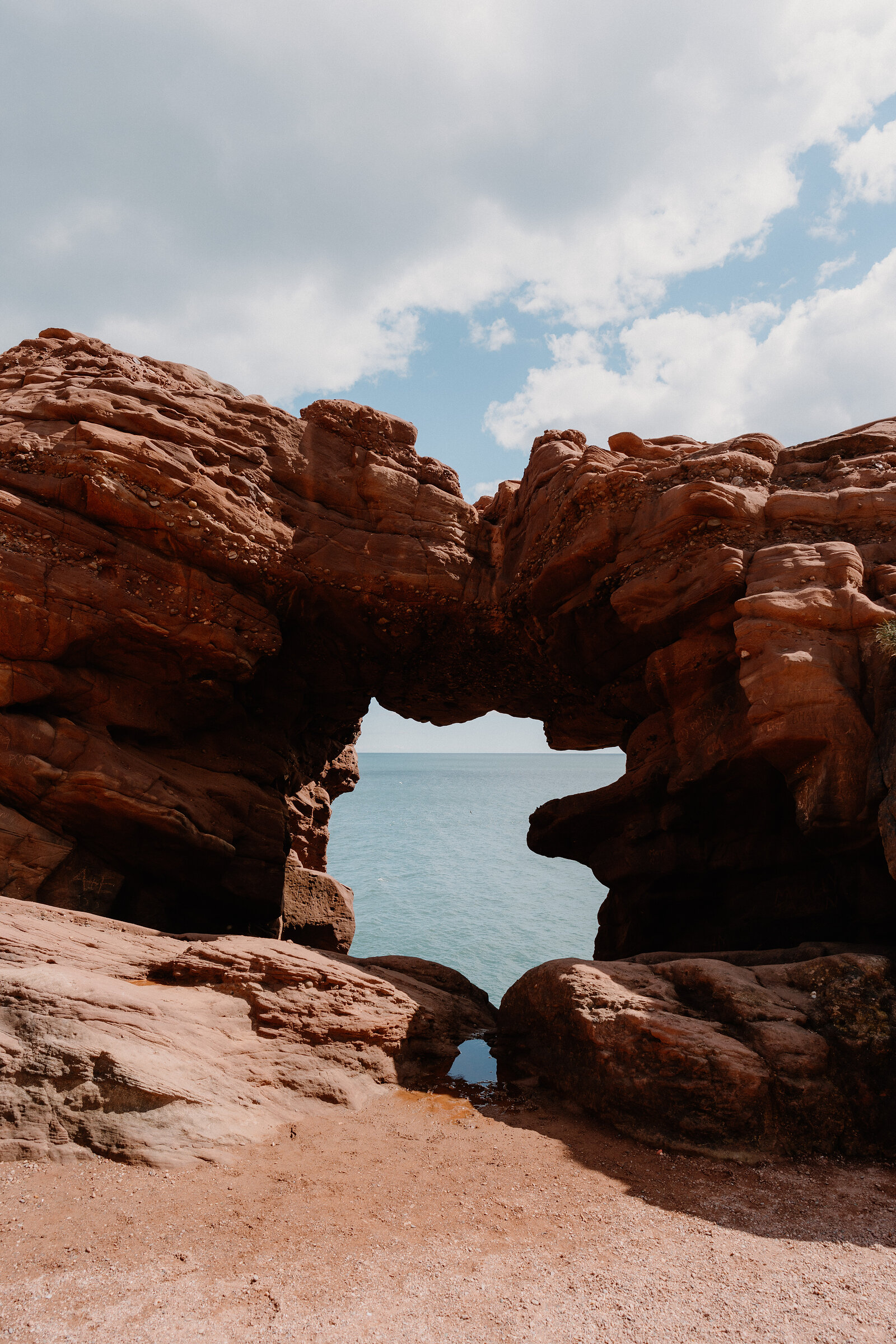 A red sandstone sea arch frames a view of open sea, layered rock and lichen covering the stone around it.