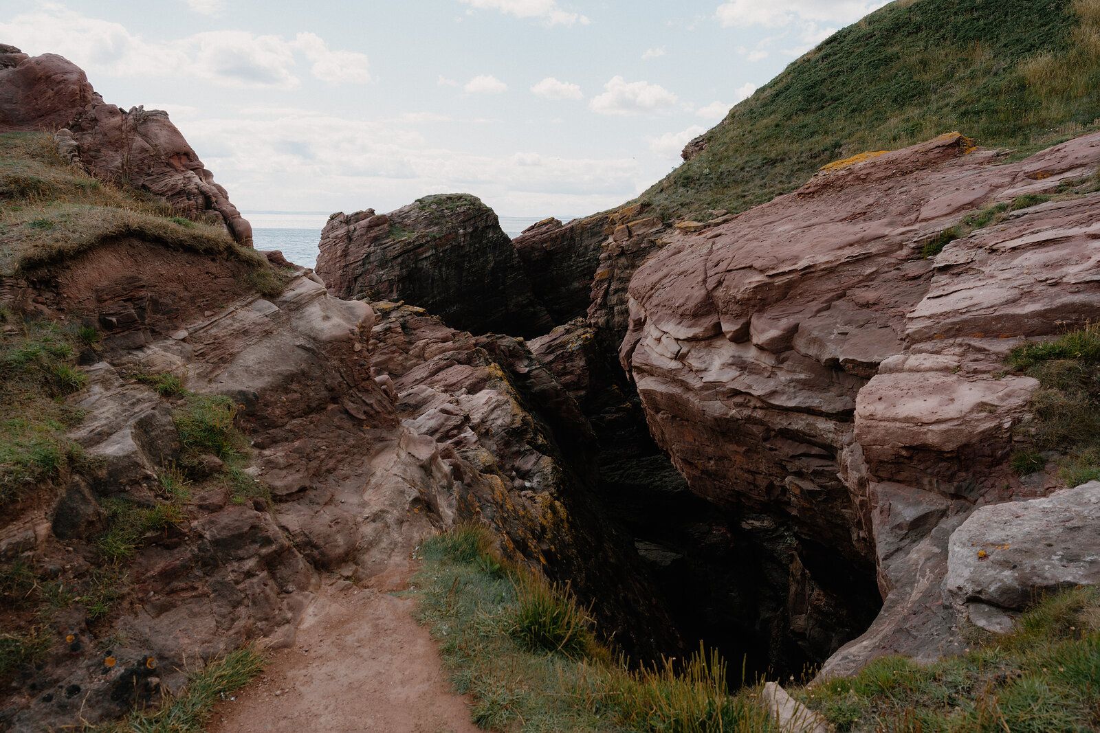A narrow path runs between red sandstone boulders split by a dark crevice, gorse and grass growing along the rock.
