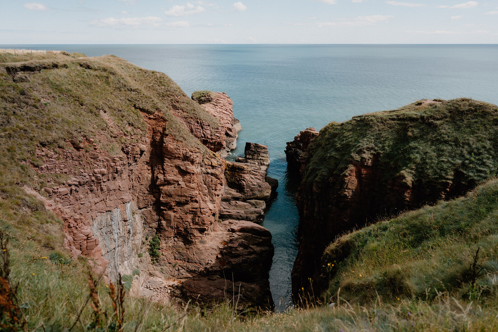 Red sandstone cliffs frame a narrow sea inlet, grass and gorse growing along the clifftop above calm blue water.
