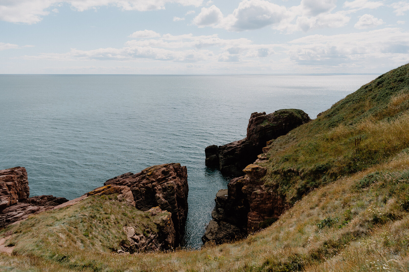 Red sandstone cliffs enclose a narrow sea channel, grass and gorse covering the clifftop above open water.
