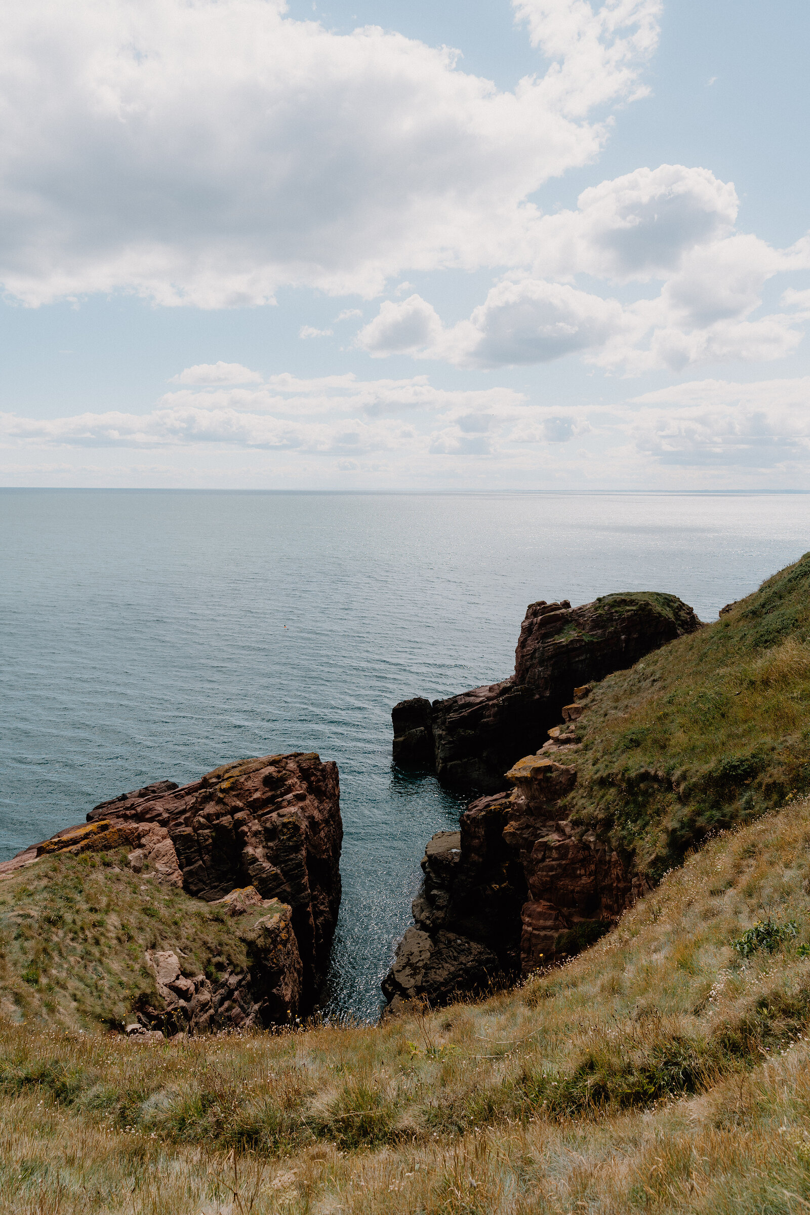Red sandstone cliffs enclose a narrow sea inlet, calm water reaching between grassy headlands under a partly clouded sky.