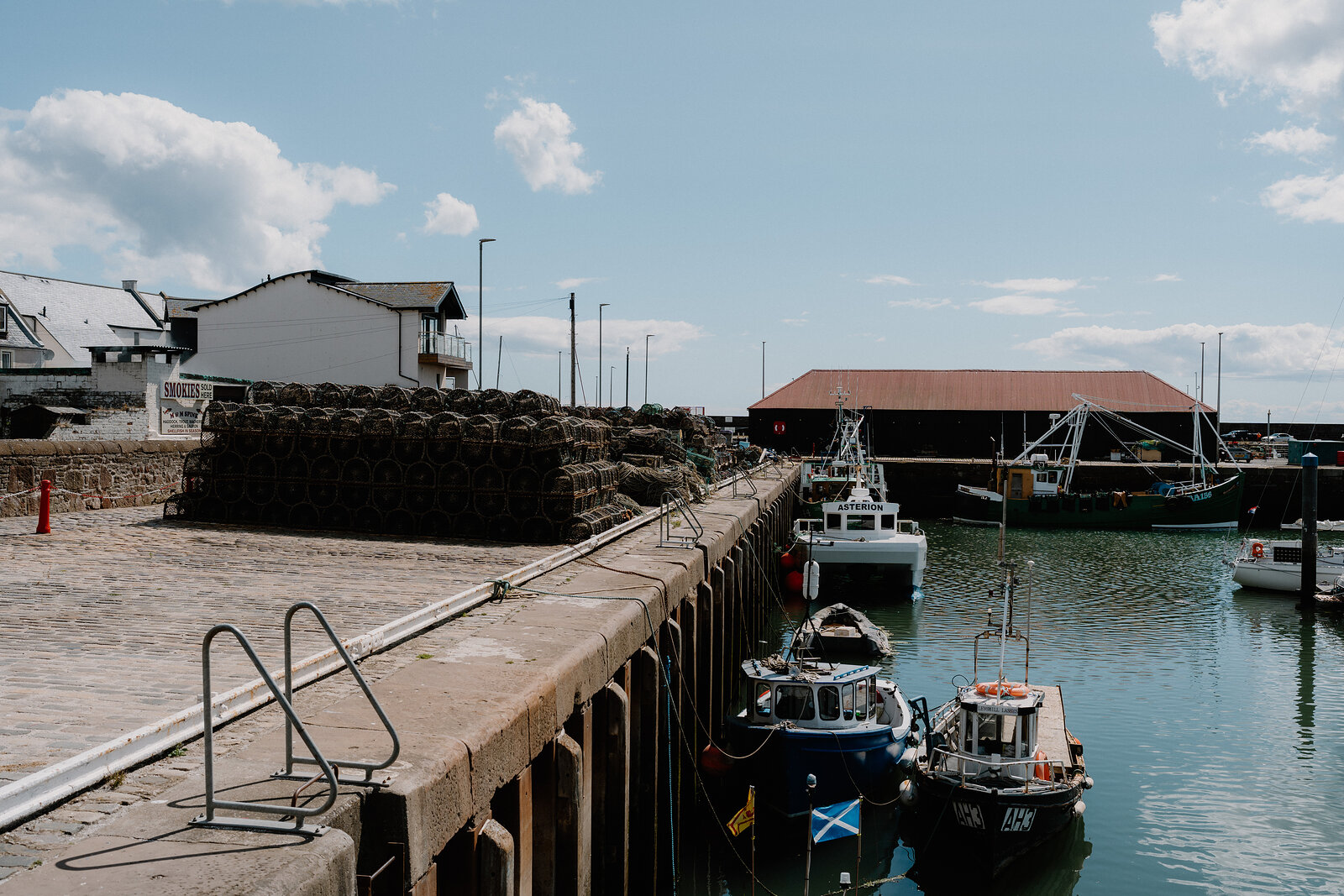 Stacked lobster creels line a stone harbour wall, fishing boats moored along the quay beside a corrugated shed.