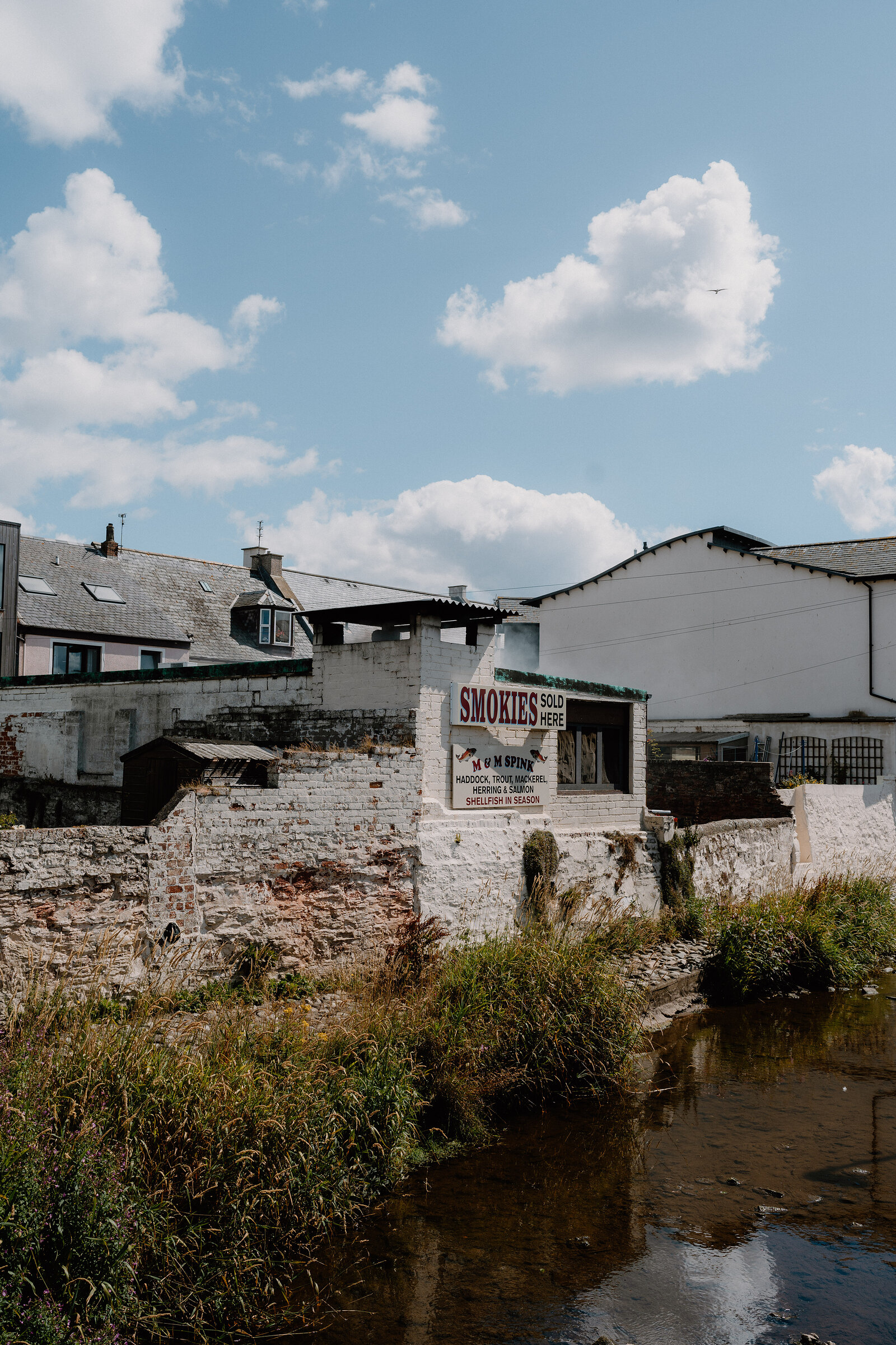 A hand-painted sign on a whitewashed harbour wall reads Smokies Sold Here, listing haddock, herring, wedding and church shellfish in season, a stream running past below.