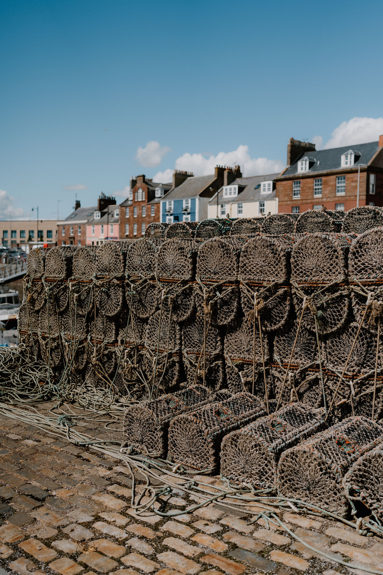 Stacked lobster creels and coiled rope line a cobbled harbour quay, colourful terraced houses rising behind under a clear sky.
