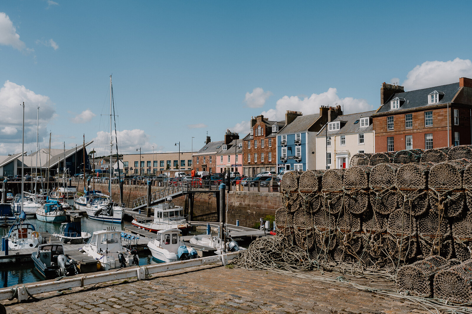 Boats and yachts fill a harbour beside stacked lobster creels, colourful terraced houses lining the quay behind.